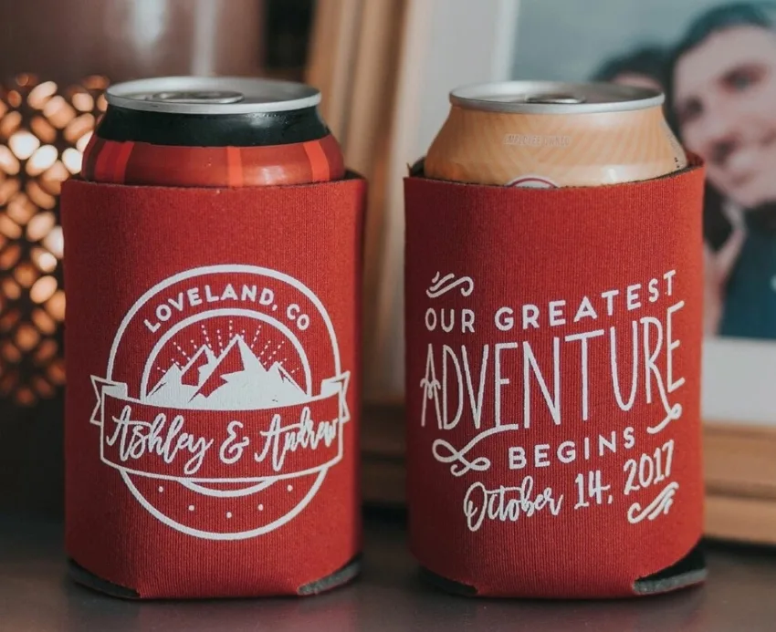 Double sided koozie with illustration of mountains on one side and the words "our greatest adventure begins" on the other side